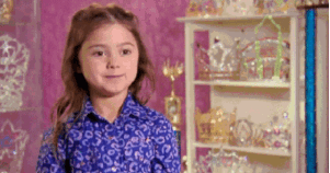 Young girl smiling with quiet confidence, surrounded by tiaras and awards.